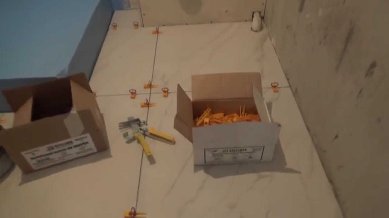 Białe płytki podłogowe z marmurkowym wzorem, ułożone z użyciem klipsów poziomujących, widoczne kartony z akcesoriami i narzędzie do poziomowania.