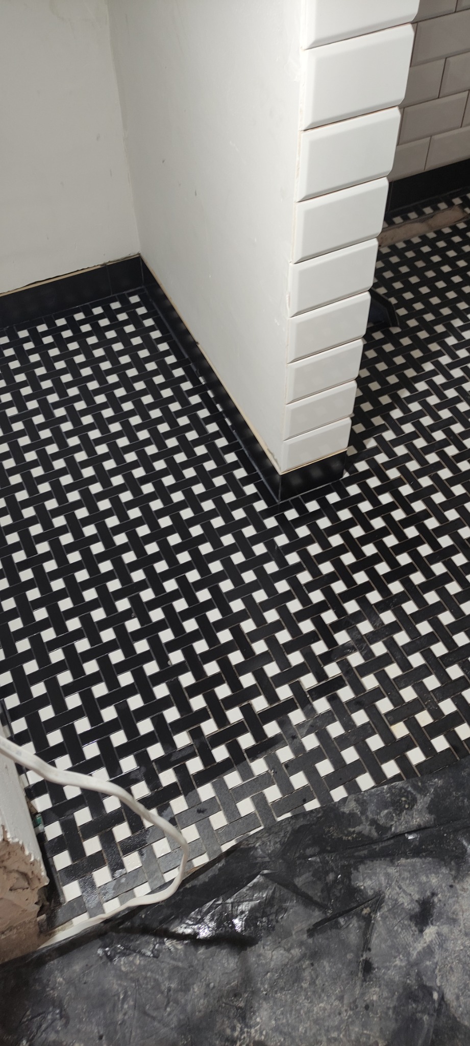 Podłoga w łazience wyłożona czarno-białą mozaiką w wzór koszykowy, obok fragmentu ściany wyłożonej białymi, prostokątnymi płytkami, widoczne ślady prac remontowych.