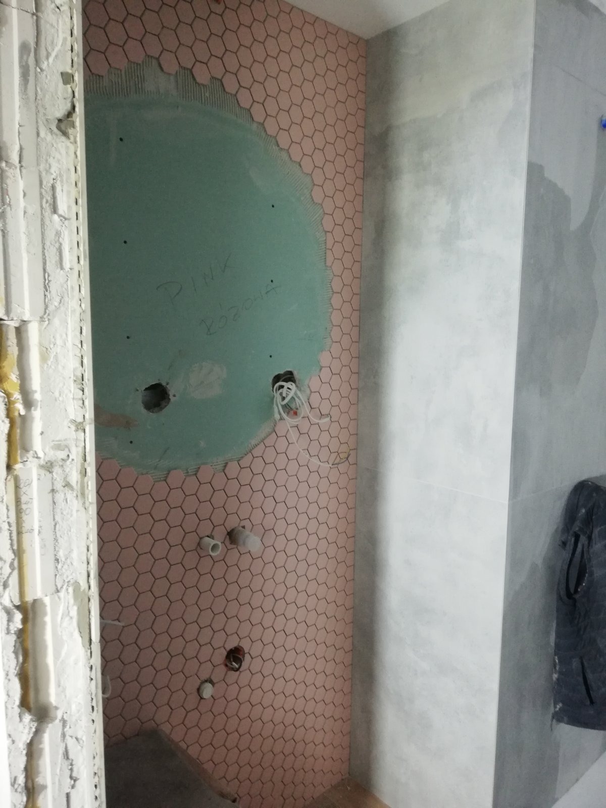 Łazienka w trakcie remontu: różowe heksagonalne płytki układane wokół okrągłego wycięcia na ścianie z płyty kartonowo-gipsowej, widoczne instalacje i fragment ściany z szarych płyt wielkoformatowych.