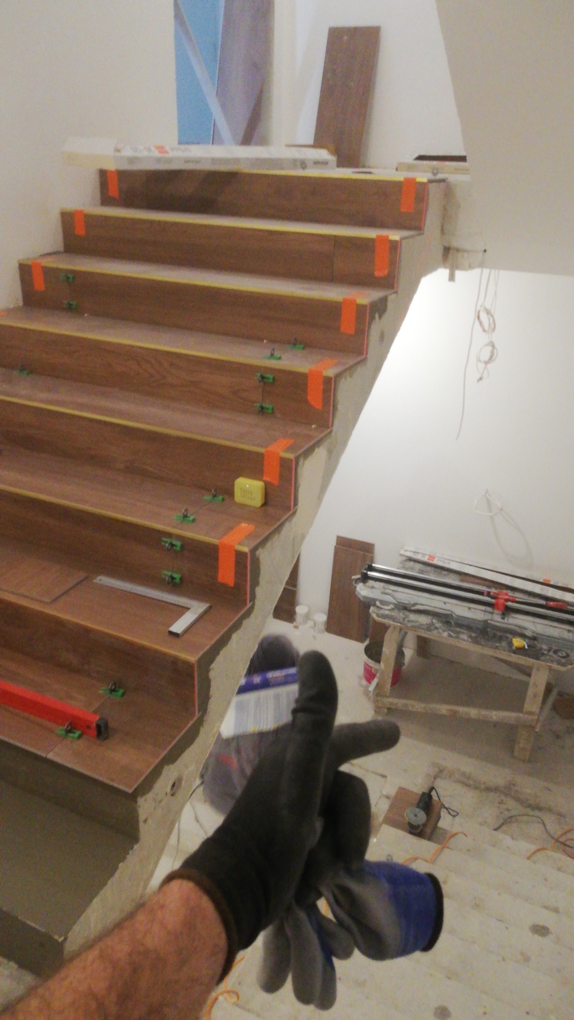 Schody w trakcie wykańczania płytkami imitującymi drewno, z pomarańczową taśmą, poziomnicą i narzędziami glazurniczymi widocznymi w tle. Ujęcie z perspektywy osoby trzymającej robocze rękawice.