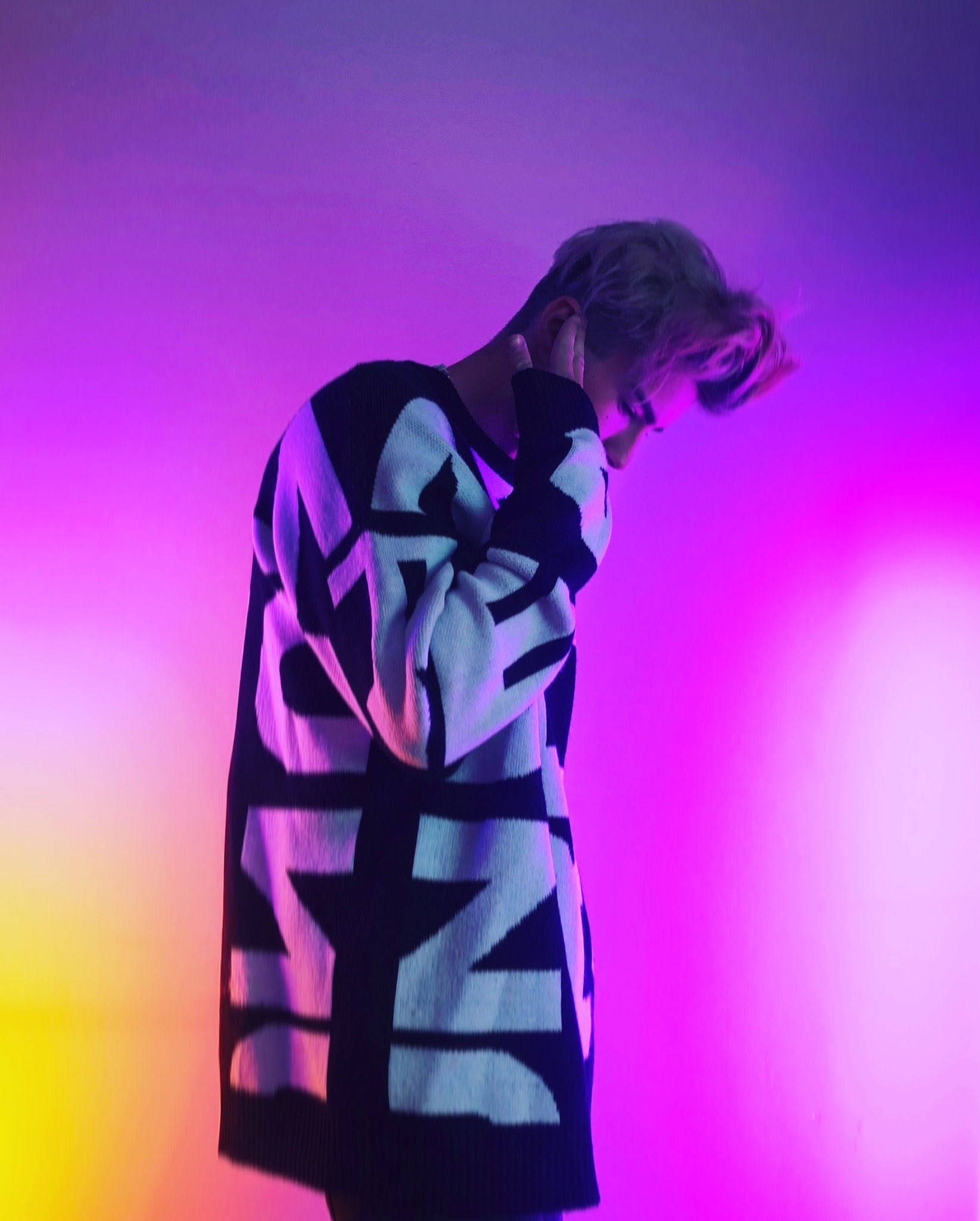 Osoba z blond włosami w czarno-białym swetrze pozuje na tle gradientu kolorów fioletu i żółci.