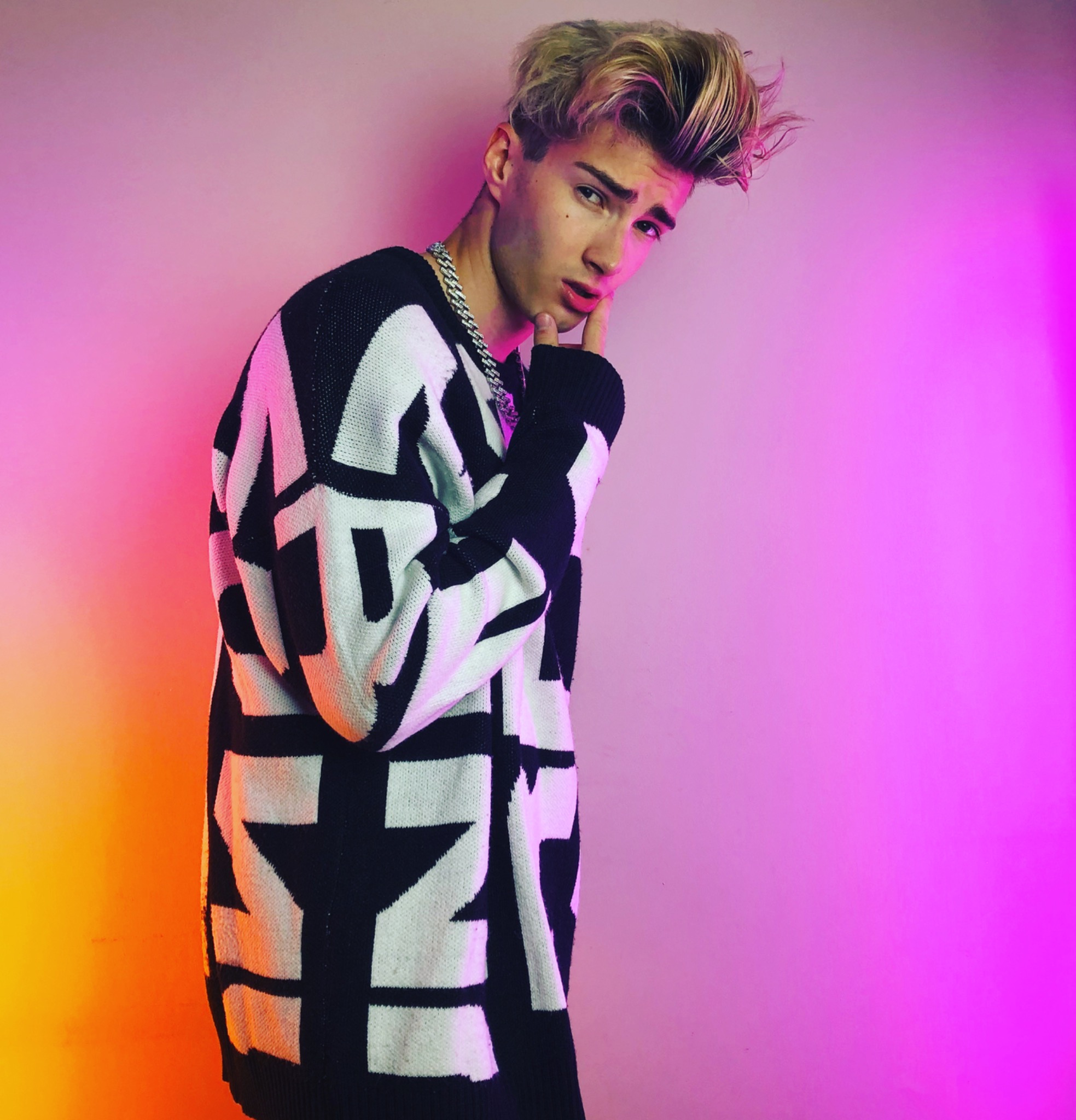 Młody mężczyzna z blond włosami w czarno-białym swetrze z geometrycznym wzorem, pozujący na różowo-pomarańczowym tle z łańcuchem na szyi i ręką podpierającą brodę.