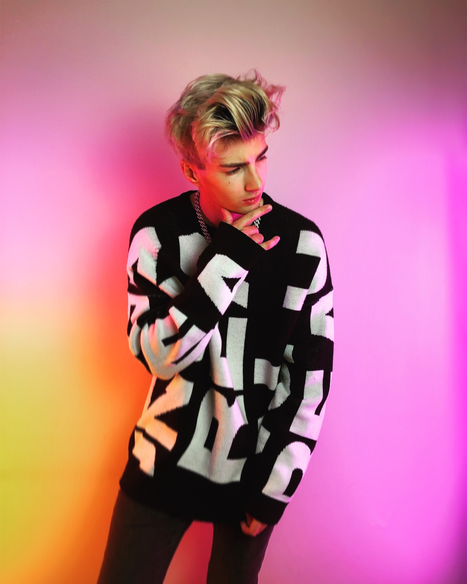 Młody mężczyzna z blond włosami, ubrany w czarno-biały sweter z geometrycznym wzorem i srebrny łańcuch, pozuje na tle z gradientem od żółtego do różowego, trzymając dłoń pod brodą.