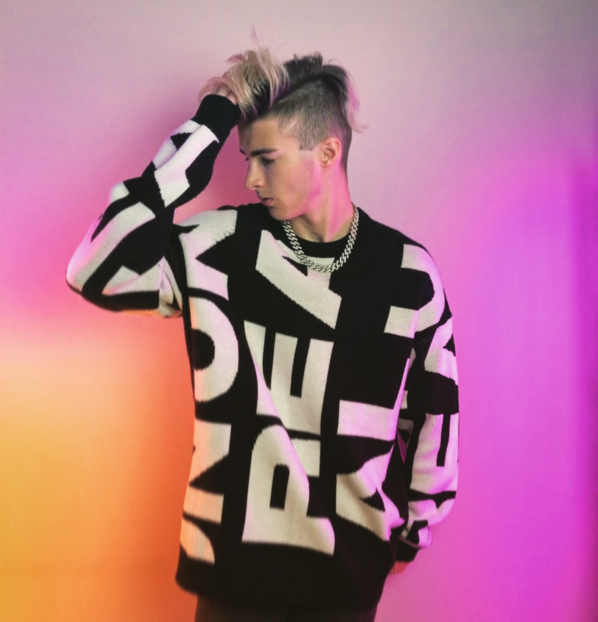 Młody mężczyzna z blond grzywką w czarno-białym swetrze z dużymi literami i srebrnym łańcuchem, pozujący na różowo-pomarańczowym tle.