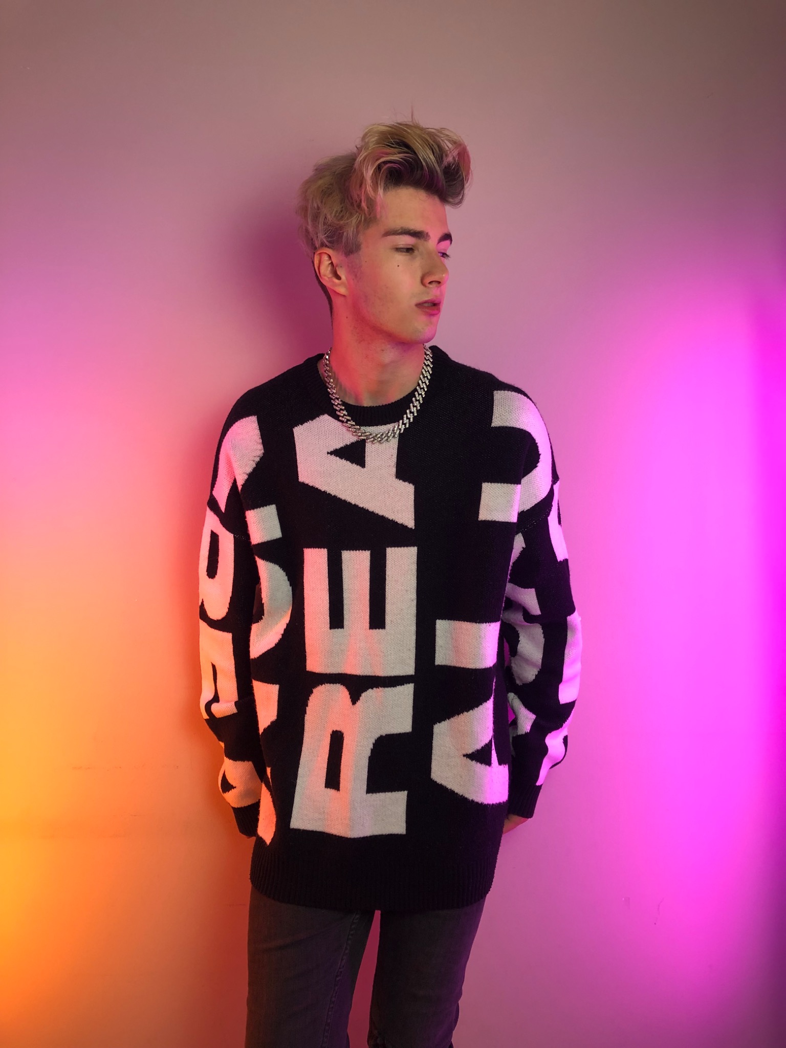 Młody mężczyzna z blond włosami w czarnym swetrze z białymi literami, pozujący na tle ściany z gradientem kolorów od różowego do pomarańczowego, z łańcuchem na szyi.