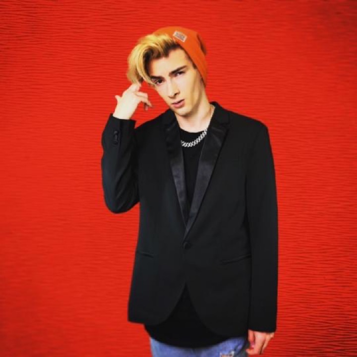 Młody mężczyzna z blond grzywką i pomarańczową czapką, ubrany w czarną marynarkę i srebrny łańcuch, pozuje na czerwonym tle, przykłada palec do skroni.