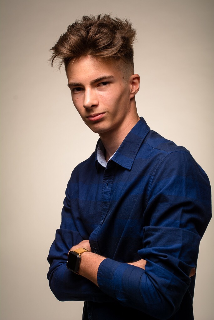 Portret młodego mężczyzny w niebieskiej koszuli z założonymi rękami, patrzącego w obiektyw, na jednolitym, jasnym tle.