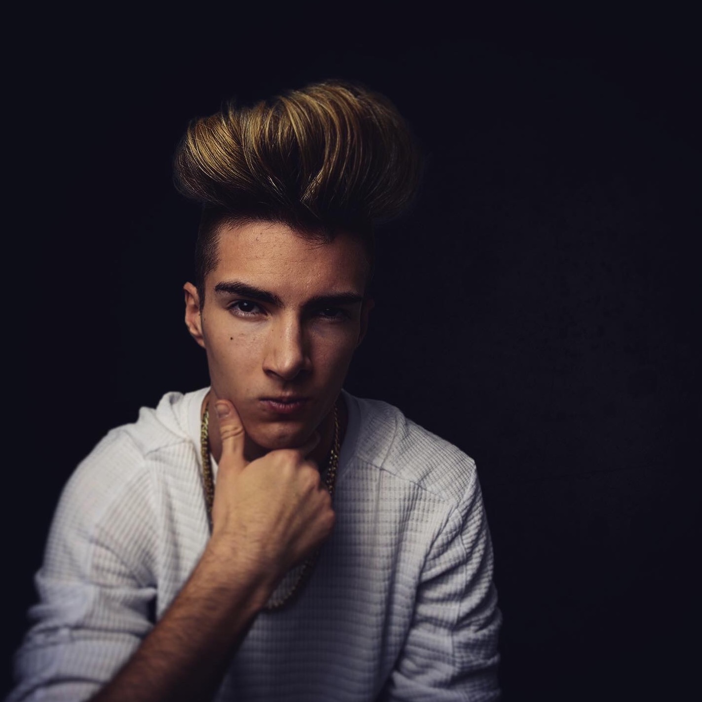 Portret młodego mężczyzny w Warszawie z charakterystyczną fryzurą, ubranego w białą koszulę i złotą biżuterię na tle ciemnego tła, z dłonią pod brodą.