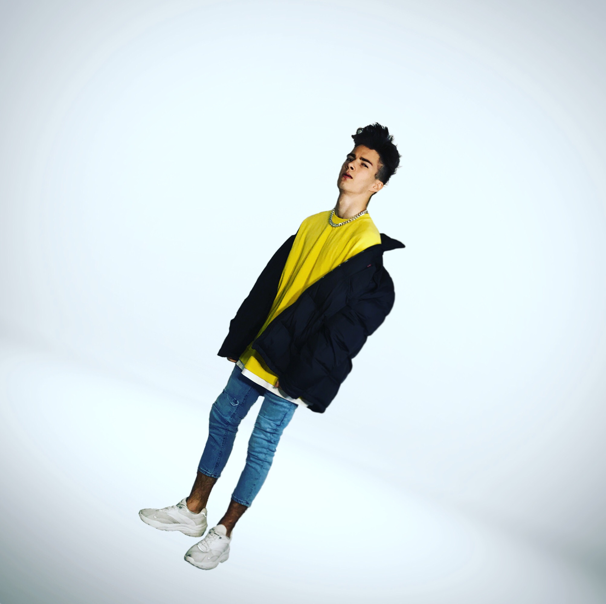 Młody mężczyzna w żółtej koszulce, dżinsach i białych sneakersach, pozuje w studio na białym tle, ubrany w czarną kurtkę, z łańcuchem na szyi, przechylony w lewo.