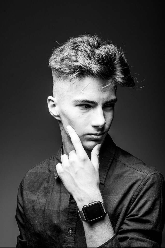 Czarno-białe portretowe ujęcie młodego mężczyzny z modną fryzurą i zegarkiem na ręce, zamyślony wzrok skierowany w dół, palec wskazujący dotyka policzka.