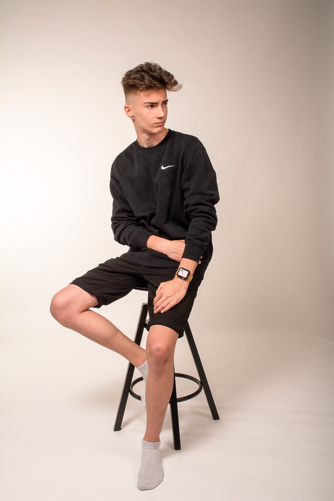 Młody mężczyzna w czarnej bluzie Nike i krótkich spodenkach siedzi na czarnym stołku barowym, spoglądając w bok. Na nadgarstku ma duży, kwadratowy zegarek.