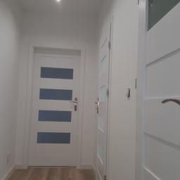 PrestigeBud - Korytarz po remoncie: widoczne trzy białe drzwi z przeszkleniami, panele podłogowe w odcieniach brązu i dwa punktowe źródła światła w suficie.