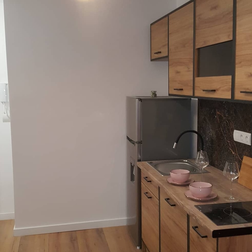 Nowoczesna kuchnia w mieszkaniu po remoncie: drewniane szafki z czarnymi obramowaniami, srebrna lodówka, marmurowy backsplash, różowe miseczki na drewnianym blacie, dwa kieliszki do wina.