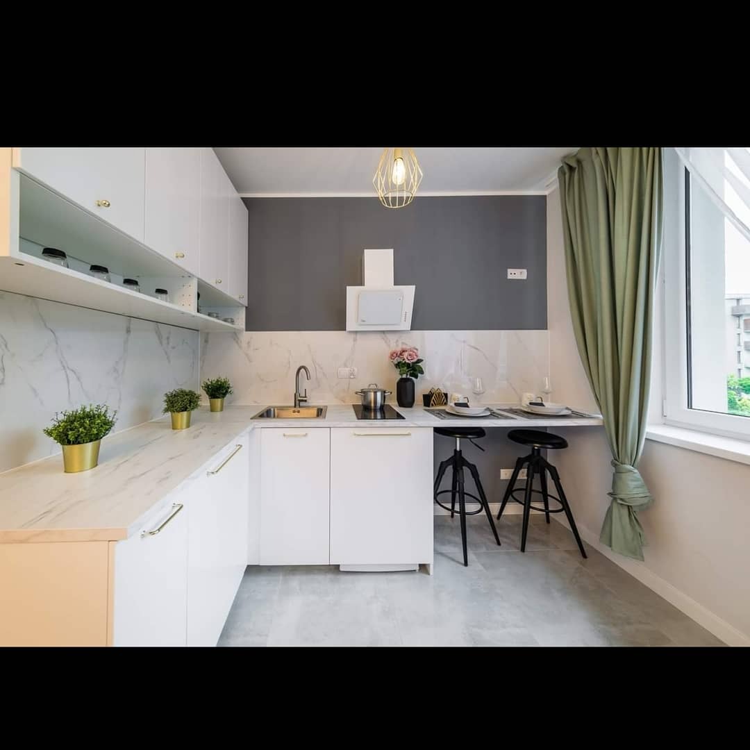 Nowoczesna kuchnia z białymi szafkami, blatem imitującym marmur, szarym akcentem na ścianie i dwoma czarnymi stołkami barowymi przy blacie, oświetlona złotą lampą wiszącą.