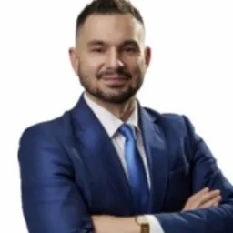 Portret mężczyzny w niebieskim garniturze i krawacie, z zegarkiem na ręce, pozującego na jasnym tle z założonymi rękami.