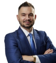 Portret mężczyzny w niebieskim garniturze i krawacie, z zegarkiem na ręce, pozującego na jasnym tle z założonymi rękami.