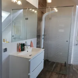 Nowoczesna łazienka na poddaszu z białą umywalką nablatową, lustrem, kabiną prysznicową typu walk-in z falistymi płytkami oraz drewnopodobną podłogą.