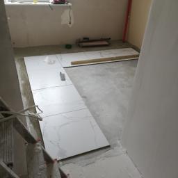 Poezja Team Marcin Gibki - Wnętrze w trakcie prac wykończeniowych: ułożone płytki podłogowe imitujące marmur, widoczna drabina, poziomica, narzędzia glazurnicze i resztki zaprawy na podłodze.