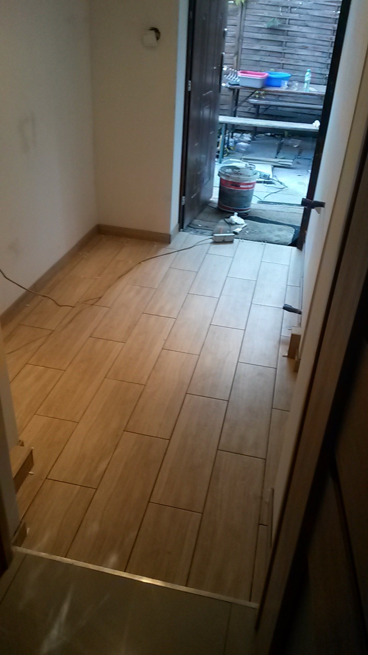 Ułożone podłużne płytki podłogowe imitujące drewno w wąskim korytarzu, widok w kierunku otwartych drzwi na zewnątrz, gdzie widać stół i akcesoria.