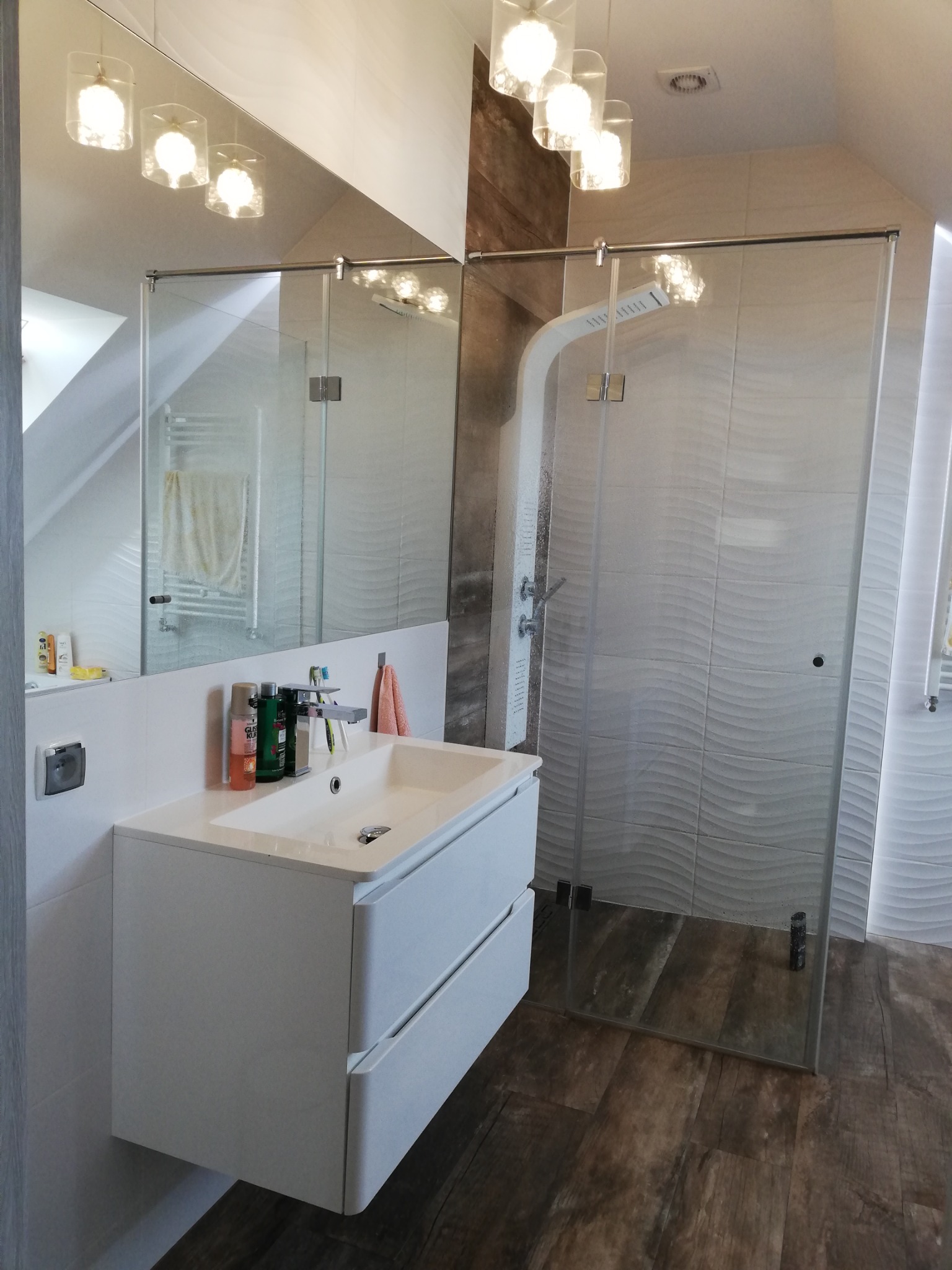 Nowoczesna łazienka na poddaszu z białą umywalką nablatową, lustrem, kabiną prysznicową typu walk-in z falistymi płytkami oraz drewnopodobną podłogą.