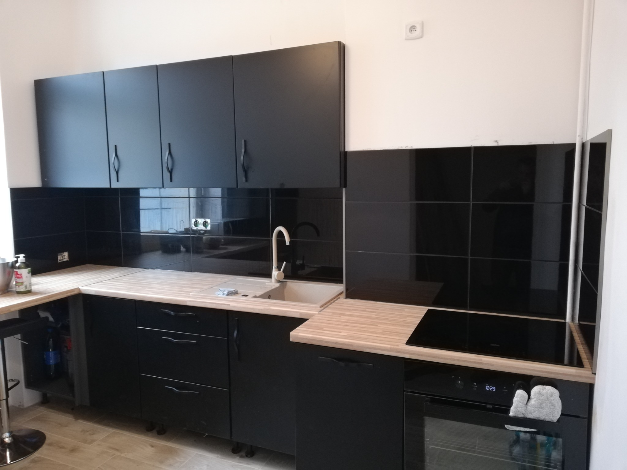 Nowoczesna kuchnia z czarnymi szafkami i drewnianymi blatami, czarne płytki na ścianie, widoczny zlew, płyta indukcyjna i piekarnik z rękawicą kuchenną.