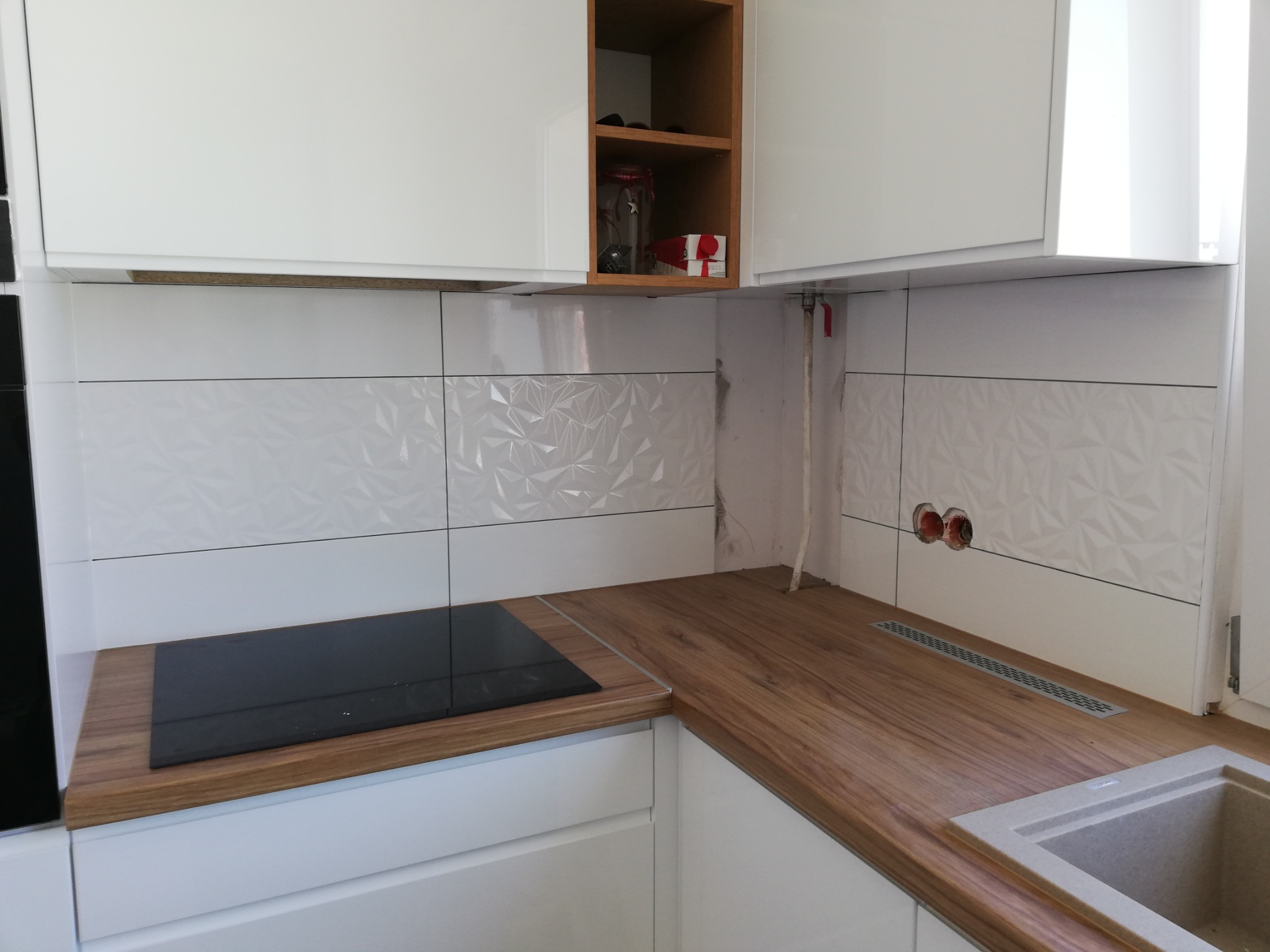 Fragment kuchni w Świdnicy w trakcie remontu: białe szafki, drewniany blat, płyta indukcyjna i ozdobne białe płytki na ścianie z widocznymi otworami na gniazdka elektryczne i rurami.