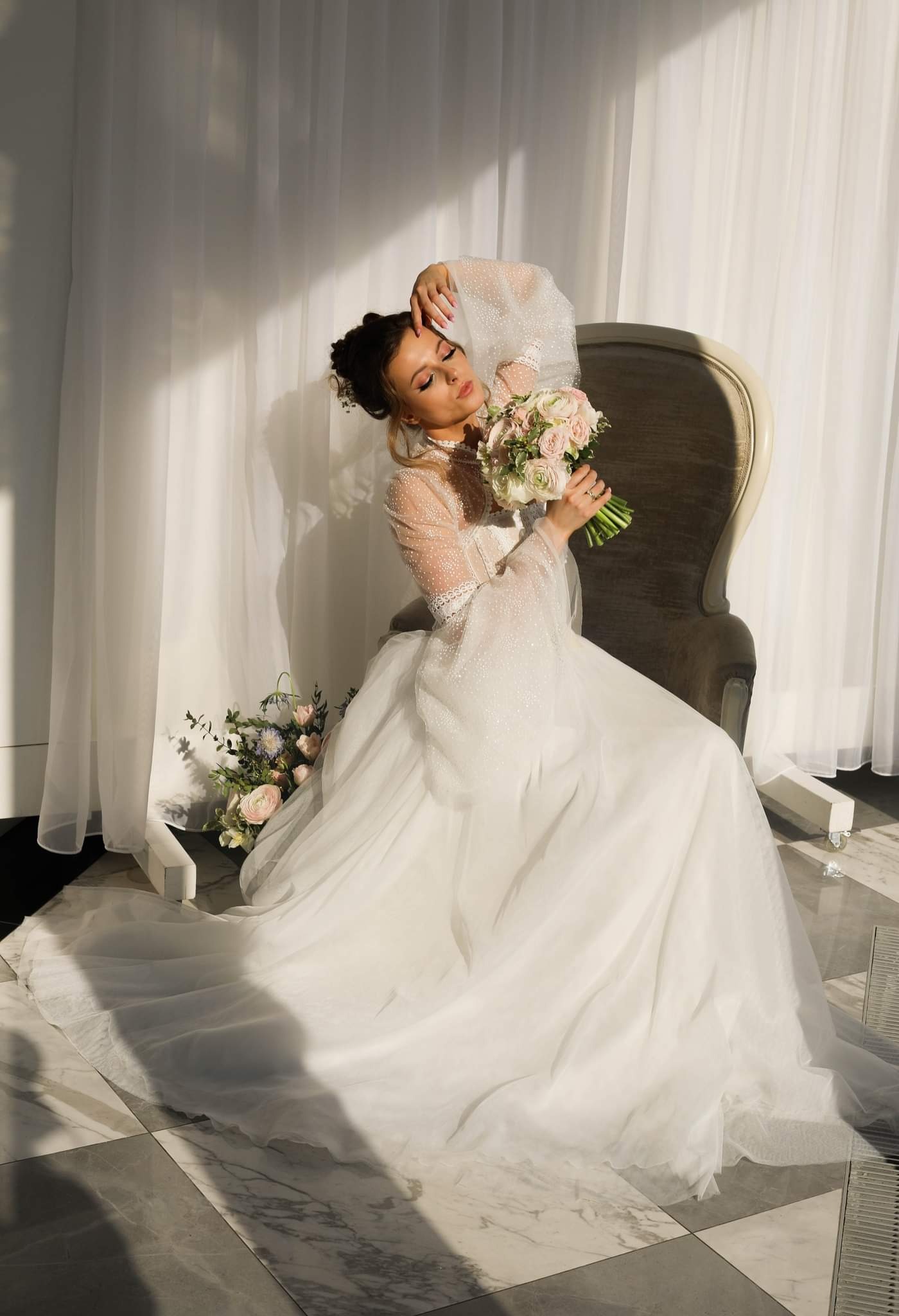 Panna młoda w białej sukni z bukietem siedzi na krześle w studio z białymi zasłonami w tle, oświetlona promieniami słońca.