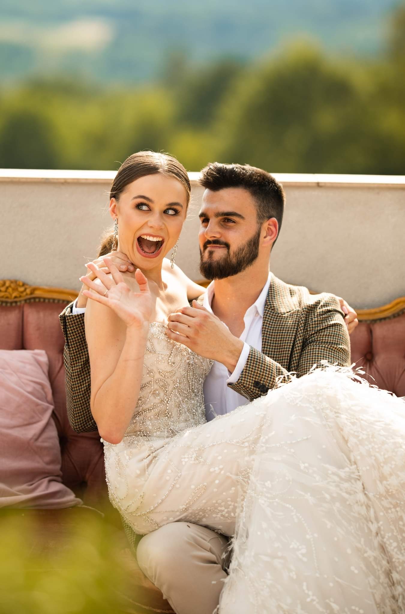 Uśmiechnięta panna młoda w bogato zdobionej sukni i pan młody w tweedowej marynarce siedzą na różowej sofie na tle zieleni.