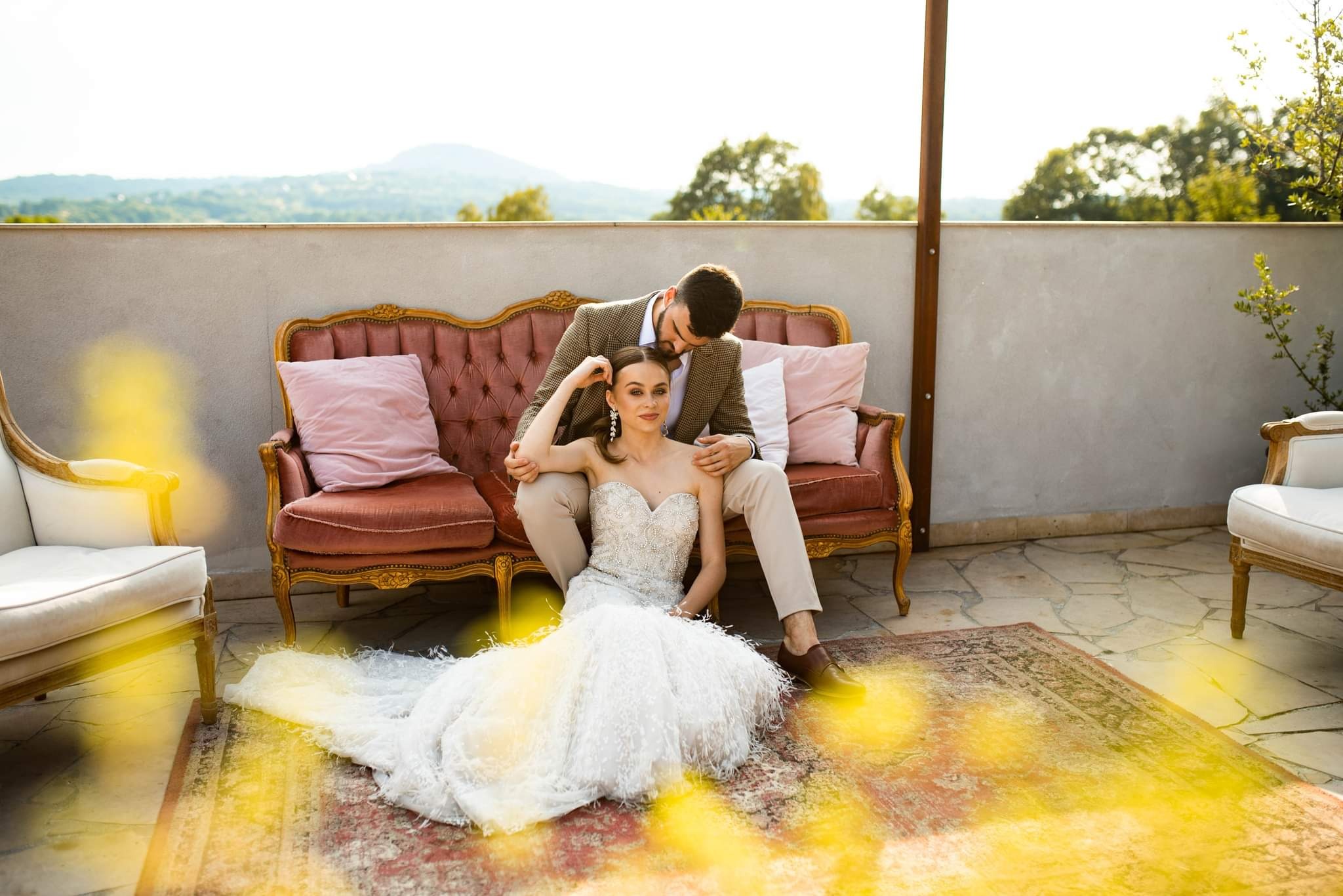 Para w strojach ślubnych pozuje na balkonie: panna młoda w sukni z piórkami siedzi na dywanie, pan młody w tweedowej marynarce siedzi na różowej sofie.
