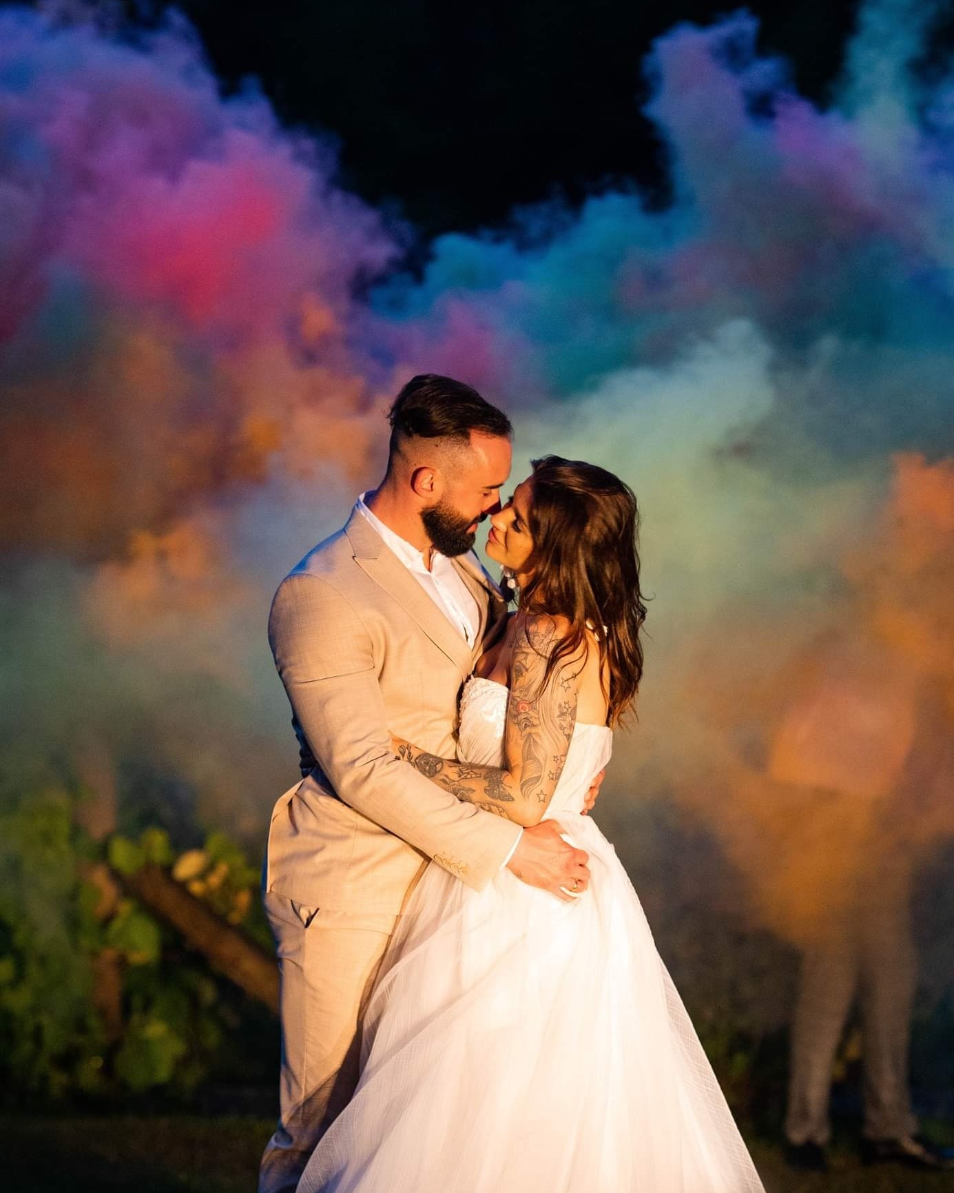 Uśmiechnięta para młoda, obejmująca się na tle kolorowego dymu podczas sesji plenerowej. Panna młoda w białej sukni bez ramiączek z tatuażami na ramionach, pan młody w beżowym garniturze, bliskość...
