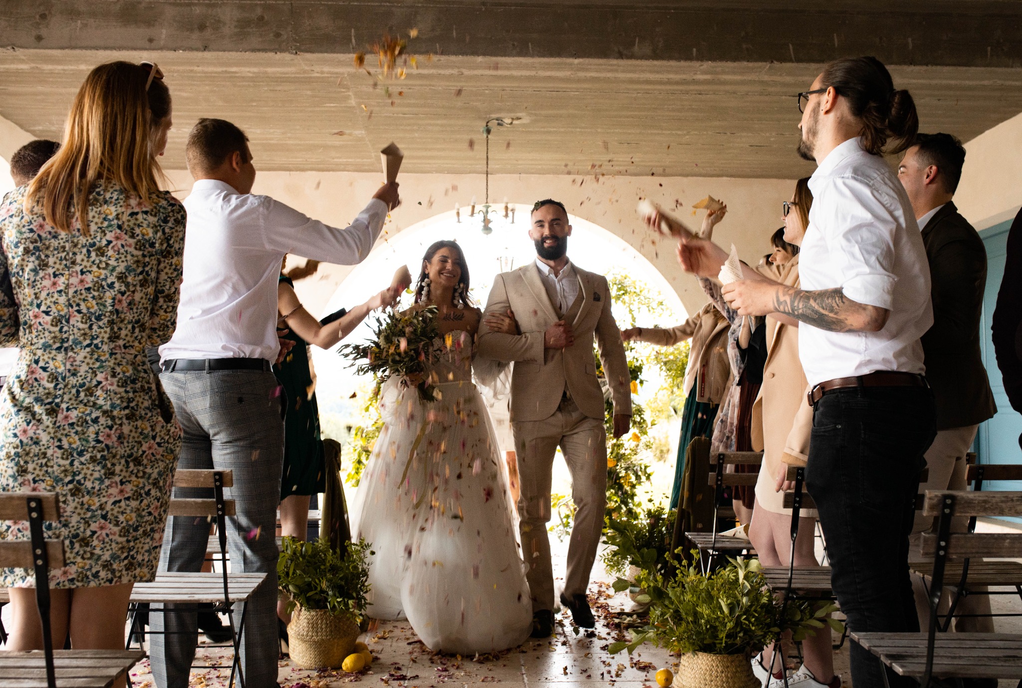 Para młoda w Lublinie przechodzi przez szpaler gości weselnych sypiących konfetti, panna młoda w sukni ozdobionej płatkami, pan młody w beżowym garniturze, rustykalny wystrój.