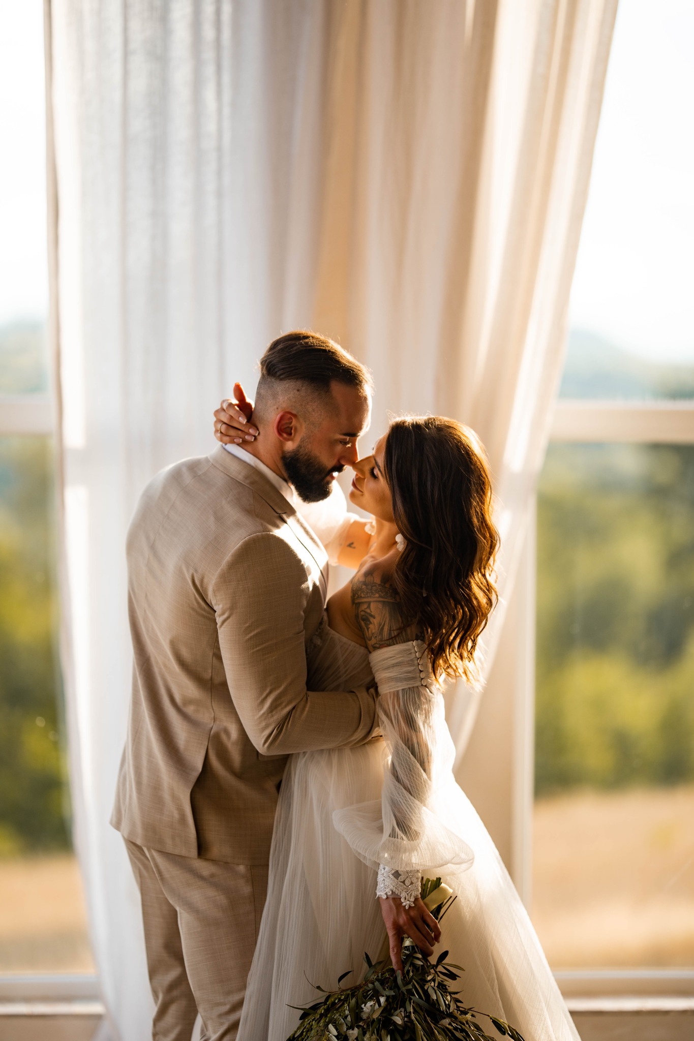 Para młoda w objęciach, panna młoda w zwiewnej sukni z bukietem, pan młody w beżowym garniturze, na tle okna z widokiem na zieleń.
