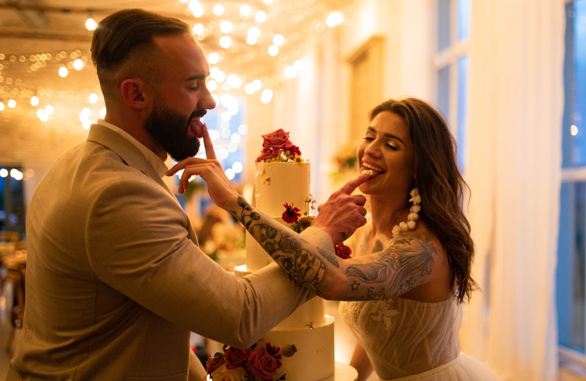 Para młoda z tatuażami, ubrana w ślubne stroje, uśmiechnięta, smakuje palcami tort weselny ozdobiony kwiatami na tle światełek.