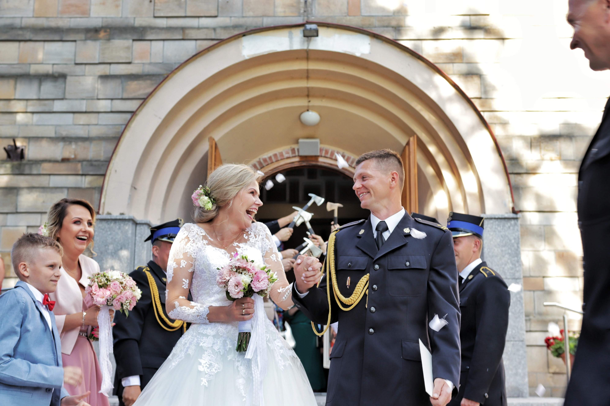 Uśmiechnięta para młoda w strojach ślubnych wychodzi z kościoła, obsypywana konfetti, w tle goście i strażacy trzymający toporki.