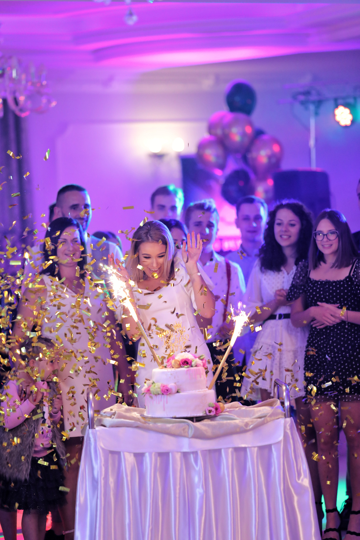 Uśmiechnięta kobieta w białej sukience, otoczona gośćmi, świętuje z tortem udekorowanym kwiatami i fontannami iskier, w deszczu złotego konfetti.