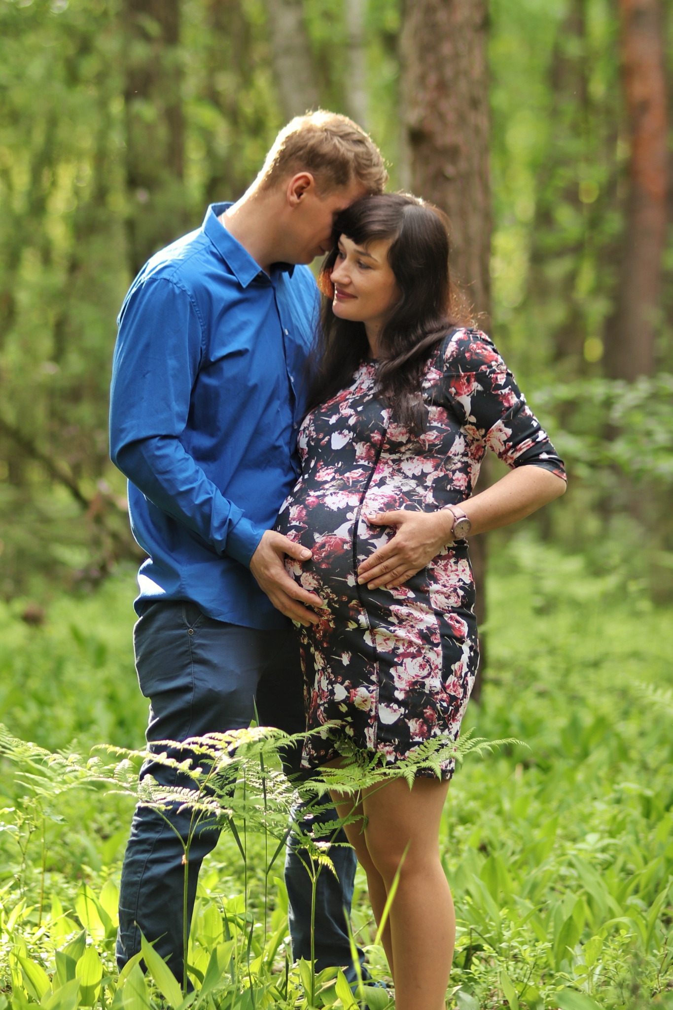 Przyszła mama w kwiecistej sukience i jej partner w niebieskiej koszuli, czule obejmujący jej brzuch na tle leśnej zieleni.