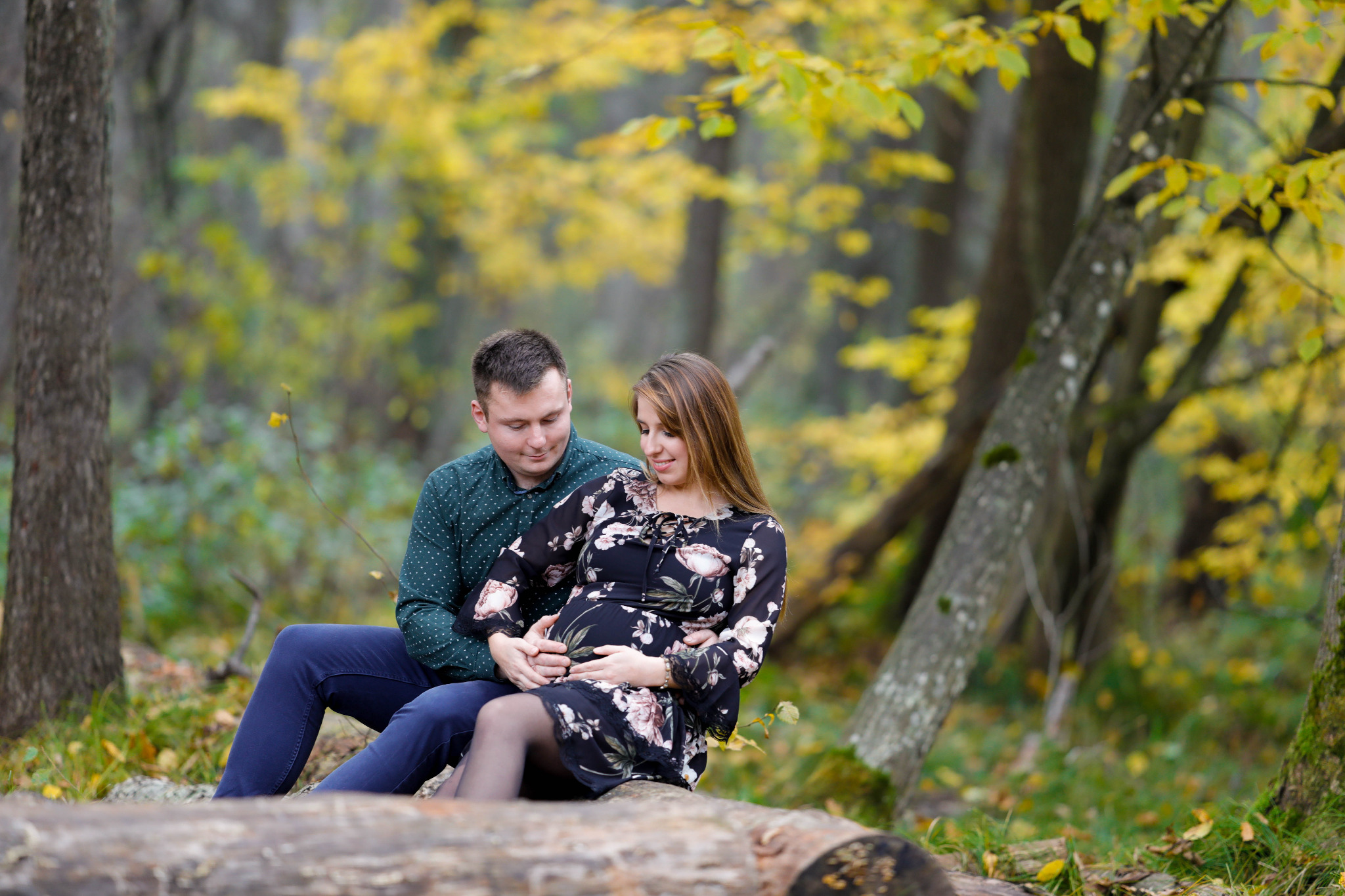 Ciężarna kobieta w kwiecistej sukience i mężczyzna w zielonej koszuli siedzą na pniu drzewa w jesiennym lesie, trzymając ręce na brzuchu.