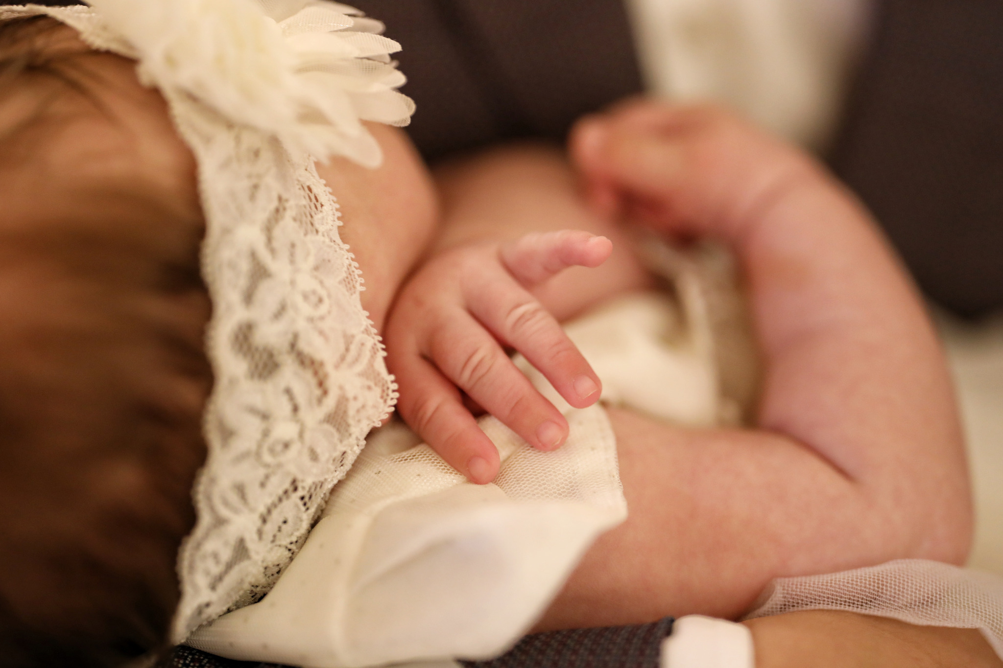 Mała dłoń noworodka spoczywa na białej, koronkowej sukience, a na głowie opaska z kwiatem, delikatne ujęcie.