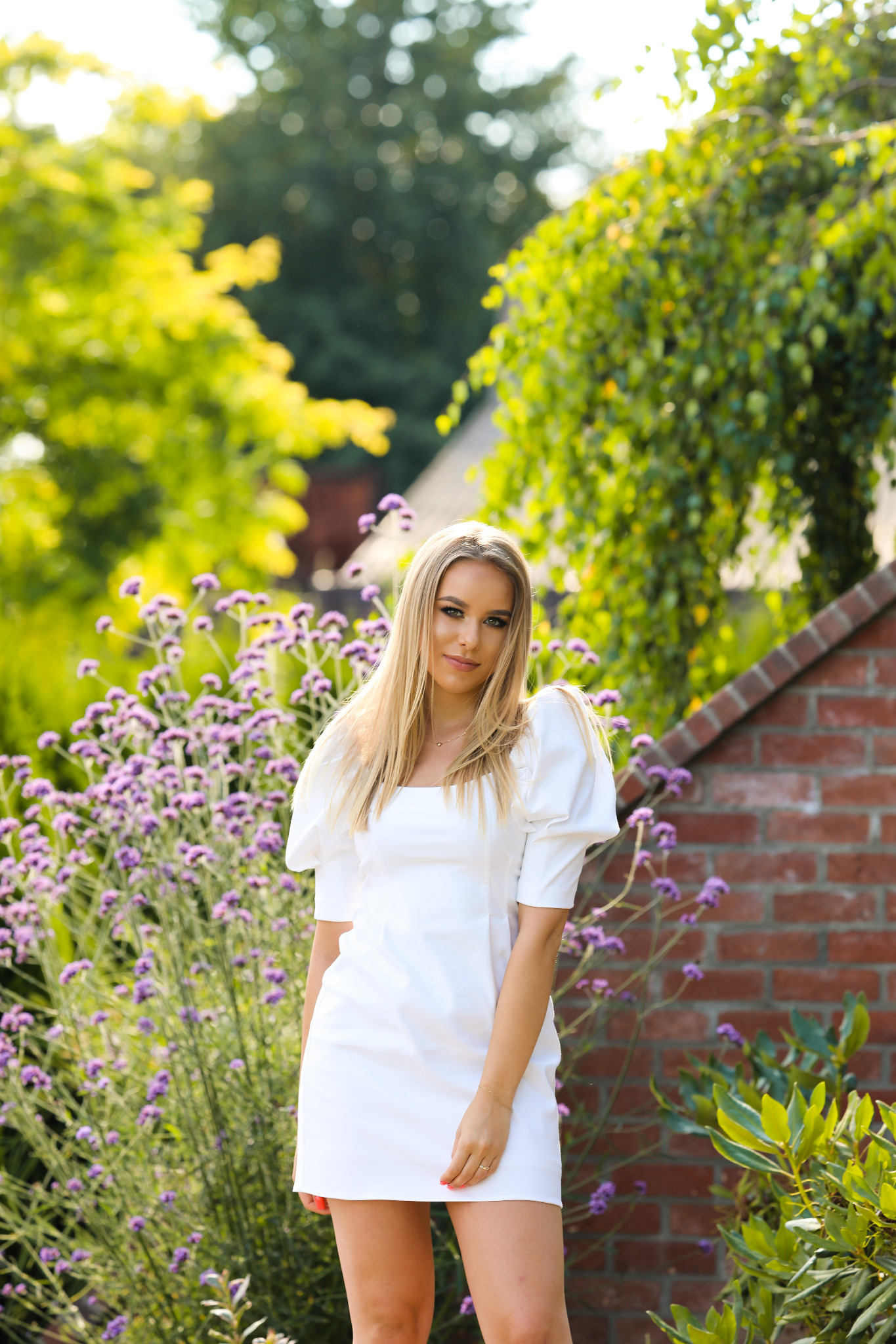 Portret młodej kobiety w białej sukience z bufiastymi rękawami, pozującej na tle kwitnących fioletowych kwiatów i ceglanego muru w słonecznym ogrodzie.