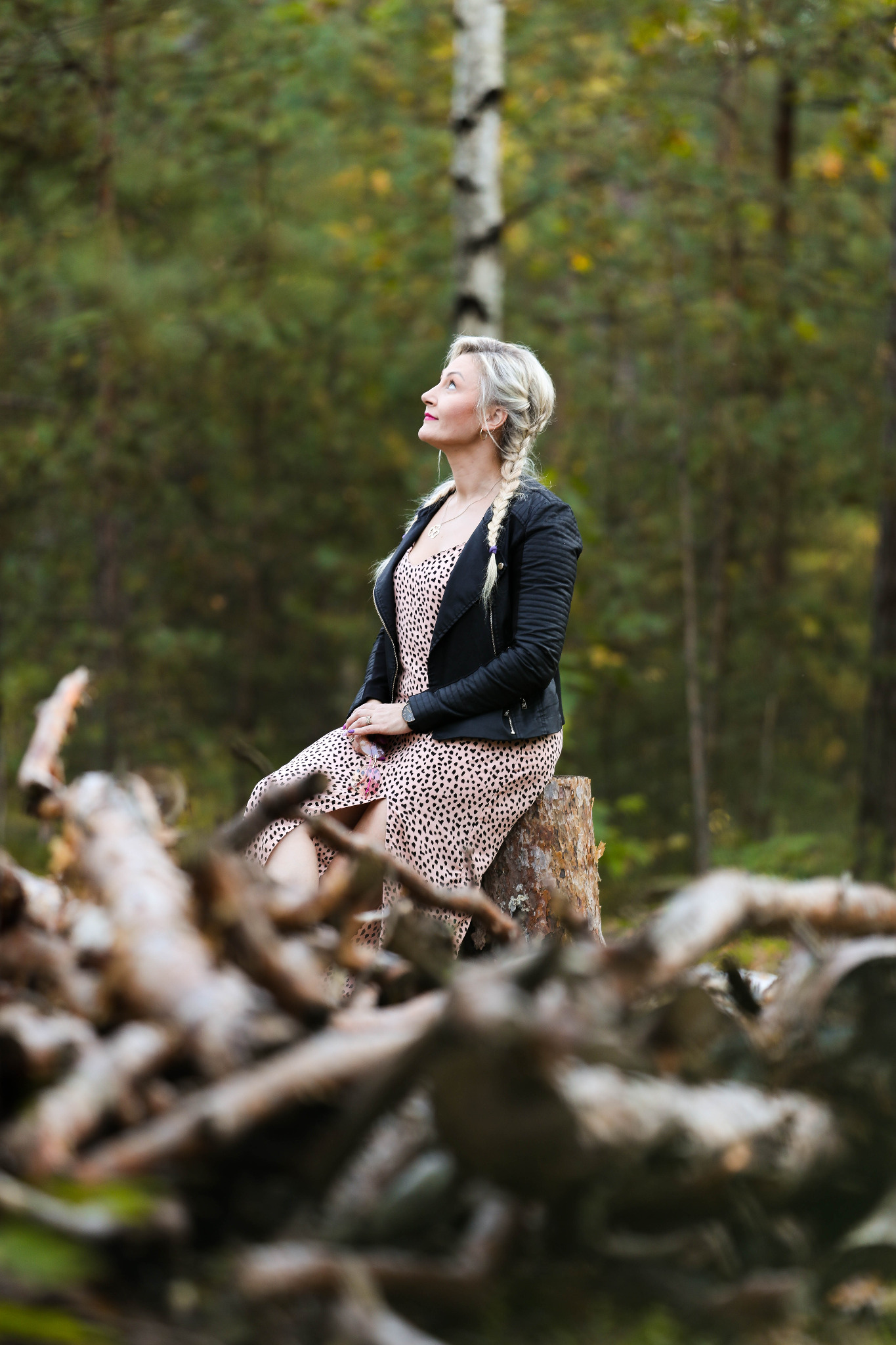 Kobieta w sukience w panterkę i czarnej kurtce siedzi na pniu drzewa w lesie, spoglądając w górę.