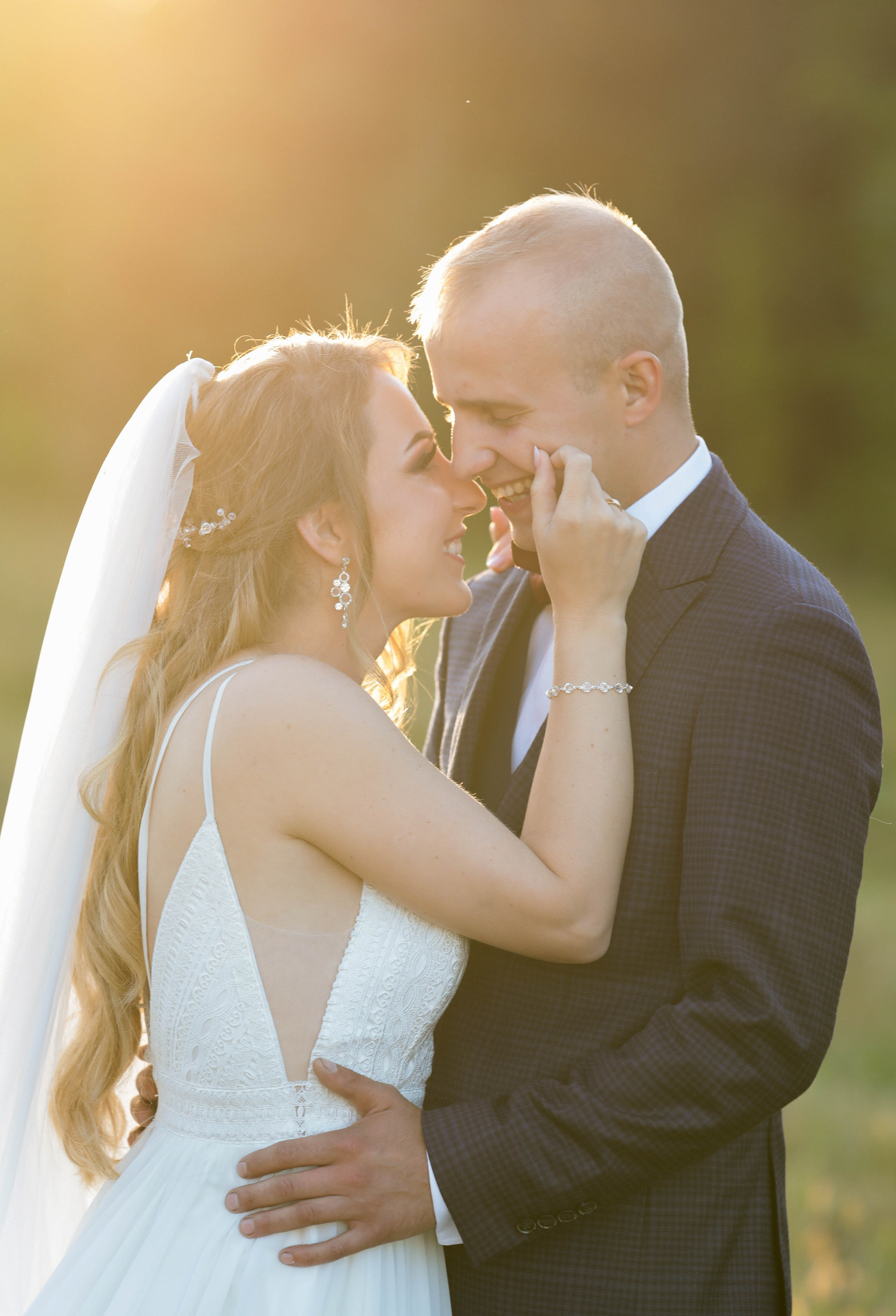 Uśmiechnięta para młoda w objęciach, panna młoda w białej sukni z koronkową górą i welonem dotyka policzka pana młodego w ciemnym garniturze w kratę, ciepłe promienie słoneczne w tle.