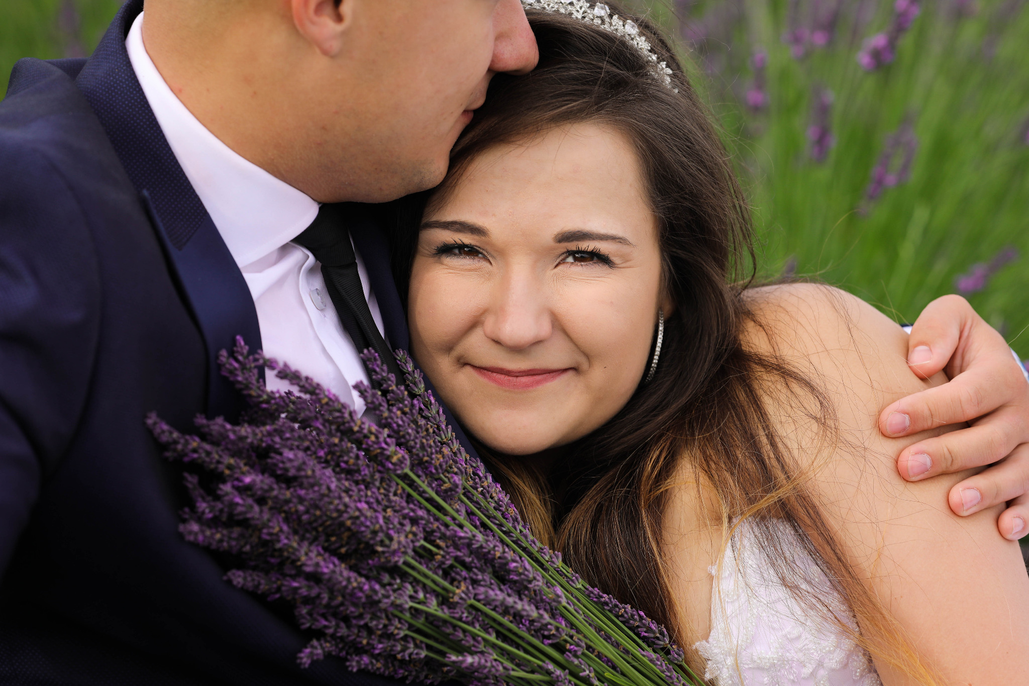 Uśmiechnięta panna młoda z bukietem lawendy w ramionach pana młodego, subtelna tiara we włosach, lawendowe pole w tle.