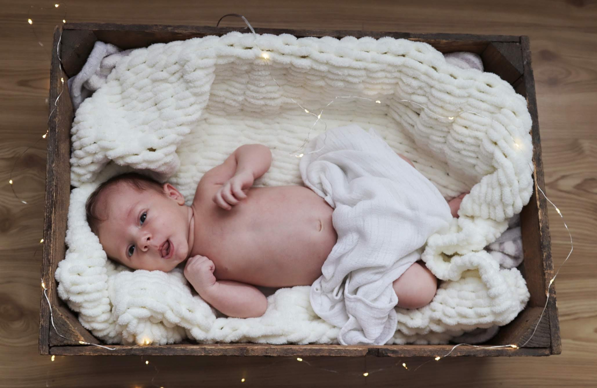 Noworodek leżący na puszystym, białym kocyku w drewnianej skrzynce, oświetlonej delikatnymi lampkami.