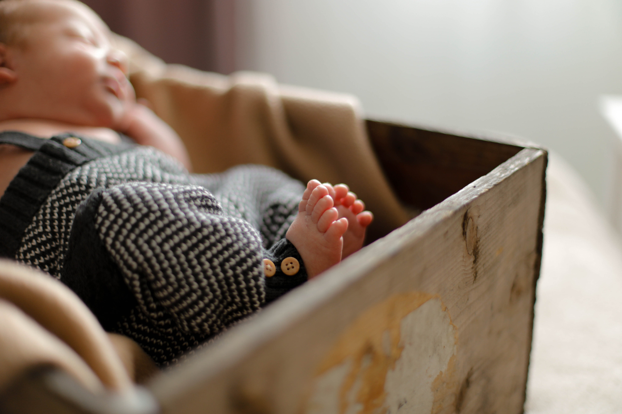 Noworodek w czarno-białym kombinezonie leży w drewnianej skrzynce, widoczne małe stopy z różowymi palcami i dwa guziki na ubranku.