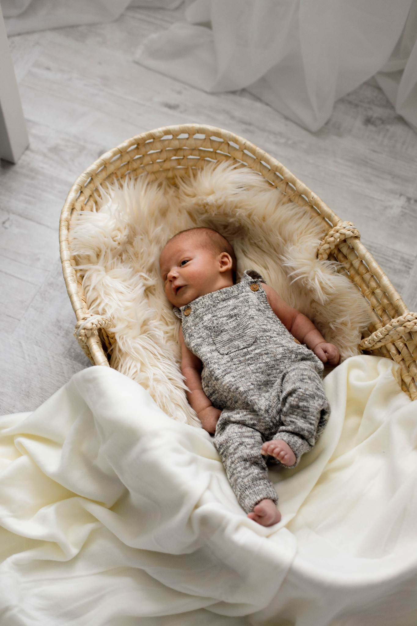 Noworodek w szarym kombinezonie leży w wiklinowym koszu wyłożonym kremowym futerkiem i białym materiałem, na tle szarej podłogi.