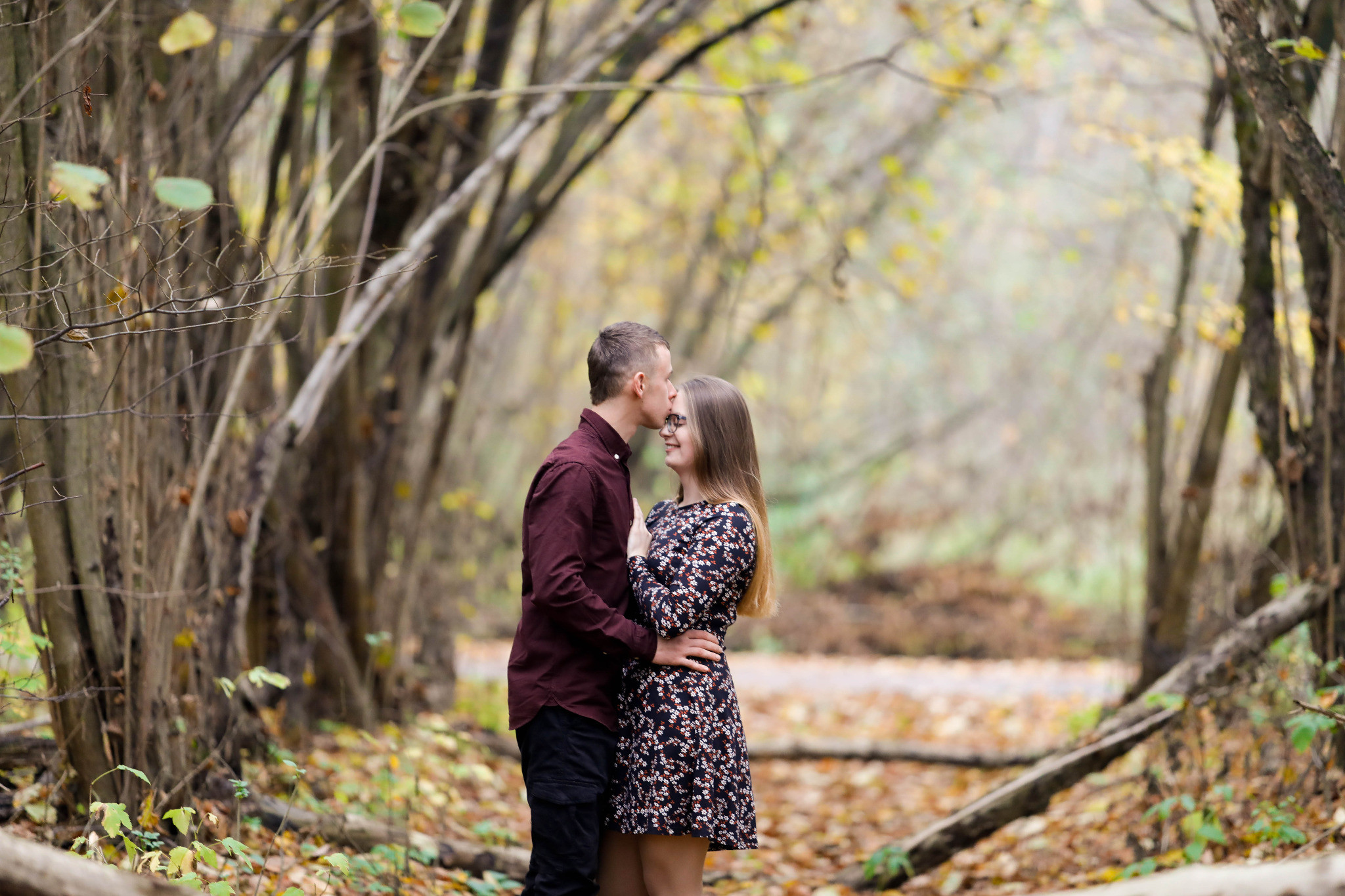 Młoda para w jesiennym lesie: mężczyzna całuje kobietę w czoło, oboje uśmiechnięci, ona w okularach i kwiecistej sukience, on w bordowej koszuli, drzewa tworzą naturalny łuk.