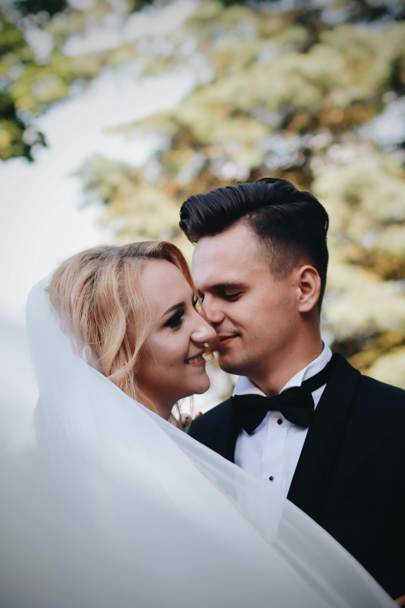 Uśmiechnięta panna młoda z blond włosami pod welonem i pan młody w czarnym smokingu z muchą, zbliżają się do pocałunku na tle rozmytych drzew.