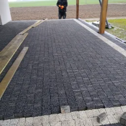 Układanie kostki brukowej na tarasie, widoczny pracownik w oddali, deski stabilizujące i niezakończony fragment z jasną kostką po bokach.