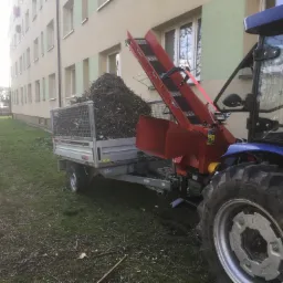 Przyczepa wypełniona pociętymi gałęziami, załadowywana za pomocą rębaka z podajnikiem, traktor w tle, teren zielony i budynek mieszkalny.