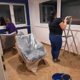 Kobieta na drabinie myje okno w nowym mieszkaniu, krzesła zabezpieczone folią, widoczne wiadro z wodą, druga osoba czyści okno obok.